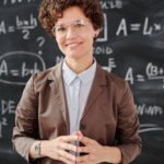 Teacher standing by a blackboard with algebra equations, smiling confidently.